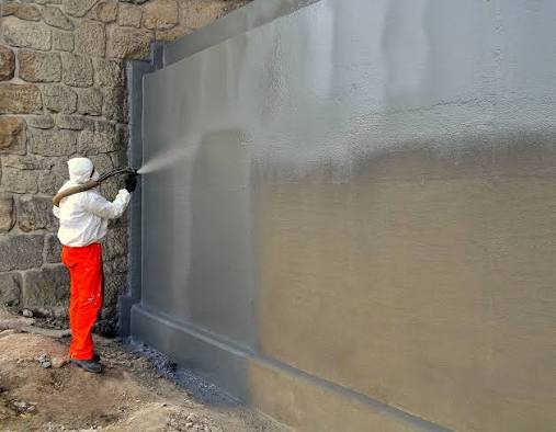 Worker spray-applying a seamless fluid waterproofing membrane onto a foundation wall